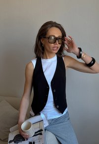 Črn gumbasti telovnik nad belo majico brez rokavov, sivi hlače, drži revijo in skodelico. Očala iz želvje lupine in naslagani črni zapestnici dopolnjujejo videz.