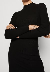 Longue robe noire côtelée avec un col montant et une silhouette ajustée, présentant des manches légèrement bouffantes et des détails en tissu texturé.