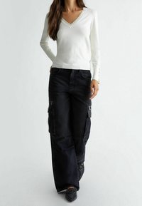 Bílý svetr s dlouhým rukávem a výstřihem do V, zdobený vlnitým lemem, kombinovaný s černými kalhotami cargo s vysokým pasem a velkými bočními kapsami.