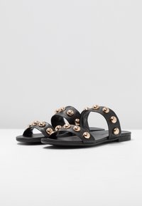 Black leather sandals with two wide straps featuring round golden studs. Flat sole, open toe design, and smooth texture.