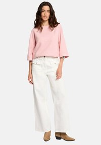 Un pull rose, ample et à manches 3/4 associé à un pantalon blanc taille haute à jambes larges. Des bottines marron à la cheville complètent la tenue.