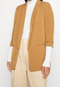 Hnědý přizpůsobený blazér s klopami, měkkou textilní strukturou a dlouhými rukávy. Nositelný přes bílý rolák; obsahuje dvě boční kapsy.