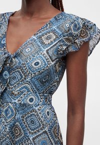 Gros plan d'une robe bleue sans manches à motifs avec un détail de volants à l'épaule portée par une personne aux cheveux tressés.
