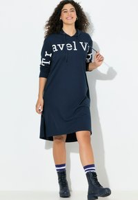 Granatowa sukienka z kapturem, z długimi rękawami, bocznymi rozcięciami i białym napisem na rękawach. Połączona z czarnymi sznurowanymi botkami i paskowanymi skarpetkami.