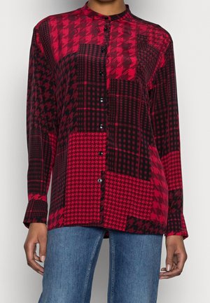 Kvinde iført en rød og sort patchwork hundetandsmønstret skjorte med knapper, lange ærmer og blå jeans.