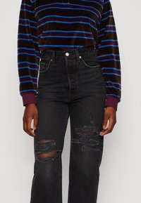 Jeans negros de talle alto con rodillas desgastadas, combinados con una camiseta de manga larga a rayas marrón y azul que cuenta con un collar y puños morados.