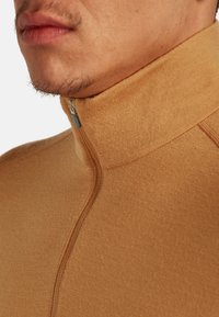 Nahaufnahme des unteren Gesichts und Nackens eines Mannes, der einen beigen Reißverschluss-Pullover mit hohem Kragen trägt, wobei die sichtbaren Nähdetails zu erkennen sind.