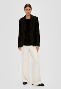 Schwarzer Blazer mit strukturierten Schultern und einem Knopfverschluss, kombiniert mit einem taillierten schwarzen Oberteil und weit geschnittenen weißen Hosen. Schwarze Schuhe.