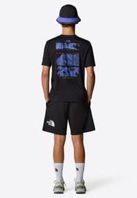 Camiseta negra con un gráfico de montañas azules, combinada con pantalones cortos negros. El conjunto incluye un sombrero tipo bucket con detalles azules y zapatillas grises.