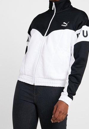 Persona con una chaqueta Puma negra y blanca con cremallera y logo en el pecho y pantalones negros, posando frente a un fondo claro y liso.