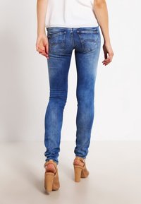 Jeans de denim azul ajustados con un ligero desgaste, diseño de cinco bolsillos y bolsillos traseros cosidos, combinados con sandalias de tacón color marrón claro.