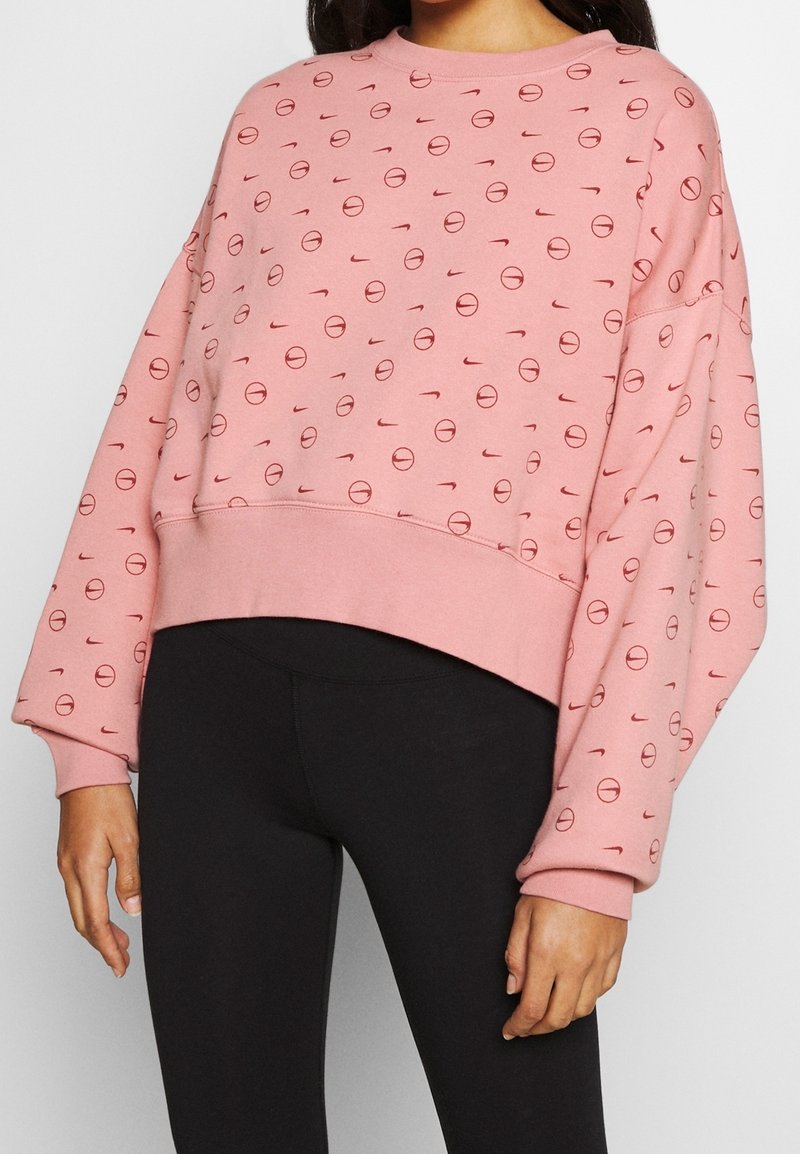 Woman wearing pink long-sleeve sweatshirt with red circular and swoosh patterns, paired with black leggings, against a plain background.