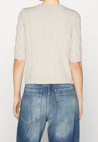 Osoba oblečená v světle béžovém pleteném svetru s krátkým rukávem a perlami, spolu s vybledlými modrými džínami s vysokým pasem, pohledem zezadu.