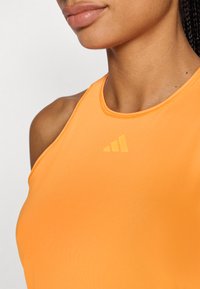 Camiseta deportiva sin mangas naranja, confeccionada con un material liso y elástico, con un cuello alto y un logo de la marca en tono similar en el pecho.