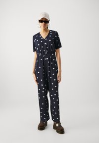 Navy polka dot jumpsuit with short sleeves, elastic waist, button front, and wide legs. Pairing brown platform shoes with a cap and sunglasses.