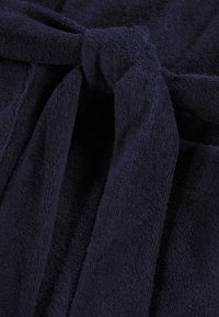 Tumši zilas kokvilnas frotē audums ar sasietu jostas detaļu un mīkstu, teksturētu virsmu. Tuva skats uz mezglu un apkārtējo audumu.