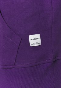 Violetti huppari, jossa on sivutasku, valkoisella neliölabelilla, jossa on "JACK&JONES"-brändäys ja ribbikuviointi helmassa.