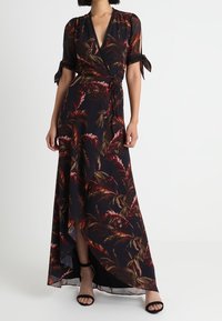 Maxi šaty z černé látky s květinovým vzorem v odstínech červené a zelené, s designem do zavazování, krátkými rukávy a bočním rozparkem. Černé sandály.