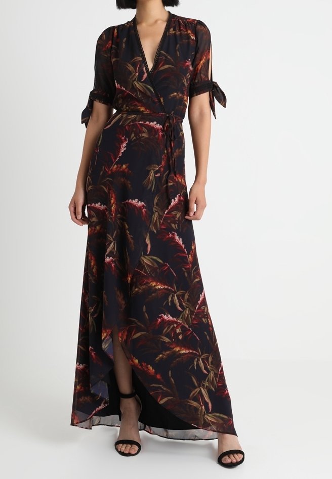 Maxi šaty z černé látky s květinovým vzorem v odstínech červené a zelené, s designem do zavazování, krátkými rukávy a bočním rozparkem. Černé sandály.