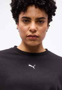 Άτομο με κοντά, σγουρά μαλλιά που φοράει μαύρο μπλουζάκι Puma με λευκό λογότυπο, στραμμένο προς τα εμπρός μπροστά σε ένα απλό ανοιχτόχρωμο φόντο.