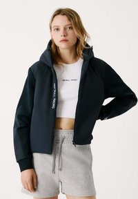 Dunkelblaues Zip-Up-Jackett mit Kapuze, versehen mit weißem Text, kombiniert mit einem weißen cropped Shirt und hellgrauen Shorts. Glatte Stoffoberfläche.