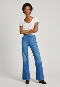 Top gris clair à manches courtes, associé à un jean flare bleu taille haute, avec une texture en denim classique et des bottines à chevilles marron foncé.