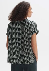 Blusa a maniche corte di colore verde scuro con un motivo geometrico bianco. Presenta un collo rotondo e una chiusura con bottone a occhio di bue sul retro.