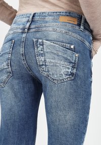 Rückansicht einer Person, die eine blaue, ausgewaschene Jeans mit gestickten Gesäßtaschen und einem hellbraunen Langarmshirt trägt.