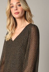 Zwarte, doorschijnende blouse met een V-hals, lange mouwen en een gouden geometrisch patroon. Lichtgewicht, met een zachte, vloeiende textuur.