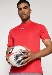 Piros Nike sportpóló rövid ujjal és kerek nyakkivágással, amelyet egy személy tart. A kezükben egy fehér focilabda fekete geometrikus mintákkal és narancssárga részlettel.