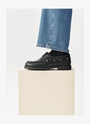Sapatos de pele pretos com bico redondo, atacadores e ilhós dourados. Sola robusta. Usados com jeans de ganga azuis que cobrem ligeiramente os sapatos.