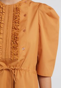 Sukienka z krótkim rękawem w ciepłym pomarańczowym kolorze, z falbanii wzdłuż przodu i guzikami. Pasek w talii tworzy dopasowaną sylwetkę.