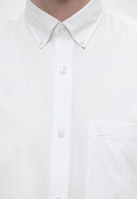 Κοντινό πλάνο ενός λευκού πουκαμίσου με κουμπωτό γιακά και τσέπη στο στήθος που φέρει μικρή κεντημένη λεπτομέρεια "L".