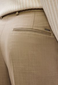 Παντελόνια σε ανοιχτό καφέ χρώμα, σε εφαρμοστή γραμμή και υφασμένα με υφής ύφασμα, με τσέπη στο πίσω μέρος και διακριτική λεπτομέρεια με λογότυπο. Κοντινή λήψη στην περιοχή της μέσης.