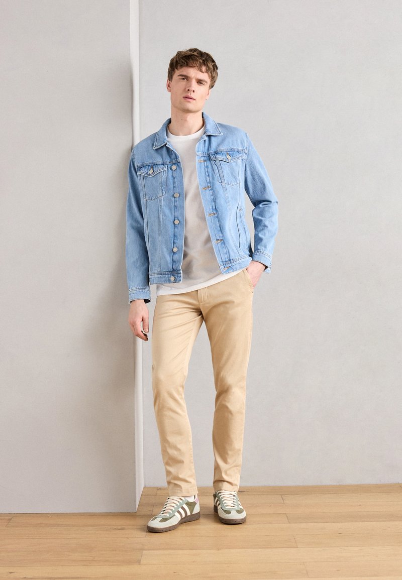Lyseblå denimjakke over en grå skjorte, beige bukser og mønstrede sneakers med grønne og hvite detaljer, stående mot en nøytral vegg.