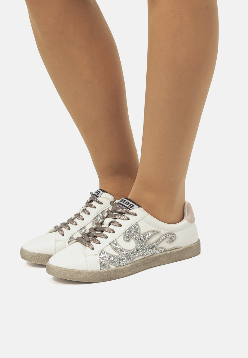 Zapatillas blancas con diseño de llamas plateadas brillantes, cordones grises y un acento rosa claro en el talón. Suela de goma con patrón texturizado.