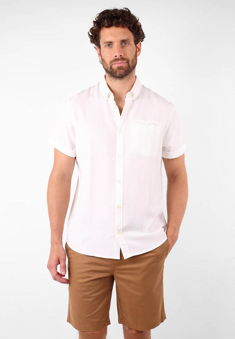 Chemise blanche à manches courtes avec une poche, associée à un short beige. Tissu lisse avec une légère texture, adapté pour une tenue décontractée.