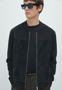 Giubbotto bomber nero con parte anteriore in suede e maniche in maglia testurizzata. Chiusura con zip argentata, abbinato a una maglietta nera a girocollo e pantaloni marroni con motivo.