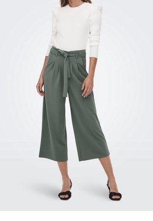 Mujer que lleva un top blanco de manga larga y pantalones cortos anchos de pierna ancha en verde oliva con cinturón atado, combinados con tacones abiertos negros.