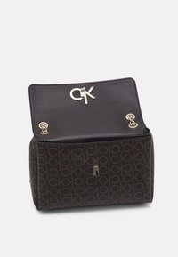 Hnědá taška s monogramem a hladkým černým koženým klopou, která má zlaté logo CK a zápinky pro bezpečné uzavření.