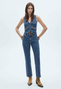 Denim top z visokim pasom in modrih kavbojkah. Top ima gumbe in kontrastno šivanje. Prilagojena zasnova s ravnimi hlačami. Rjavi gležnjarji.