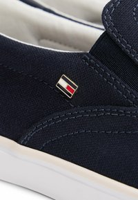Granatowy sneaker z płótna na wsuwkę z białą podeszwą i małą prostokątną plakietką z logo w kolorach czarnym, białym i czerwonym po boku.