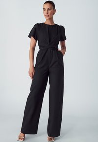 Zwarte jumpsuit met korte pofmouwen, een wikkelontwerp, wijduitlopende broekspijpen en een gestructureerde stof. Heeft een gedefinieerde taille en een minimalistische stijl.
