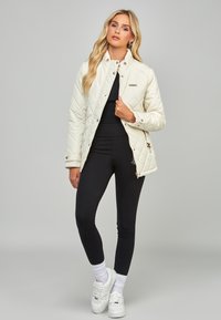 Pudret cremet jakke med ståkrave, sølv hardware og sidelommer, parret med sorte leggings og hvide sneakers.