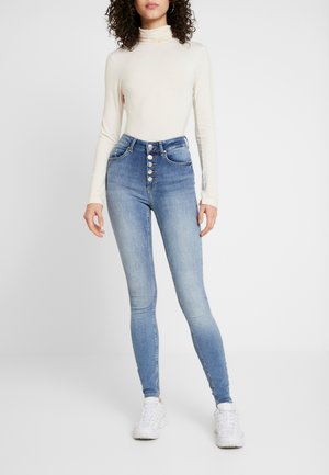 Jeans ajustados de cintura alta en azul con cierre de botones en la parte delantera, dos bolsillos frontales y una textura desgastada, combinados con una blusa de manga larga beige.