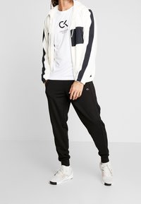 Witte en marineblauwe zip-up jas, grafisch wit T-shirt, zwarte joggers en witte sneakers met accenten. Logo details op de kleding.