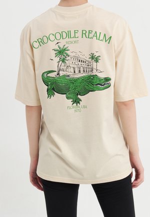 Beige T-shirt met groene afbeelding van een alligator, palmbomen, een gebouw en de tekst "Crocodile Realm Resort Florida USA 1970" op de achterkant.
