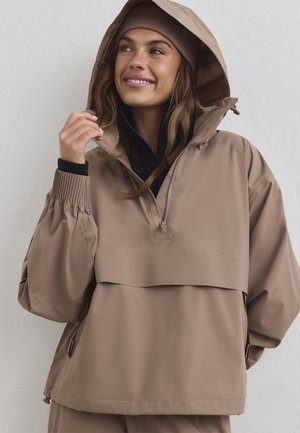 Jeune femme portant une veste à capuche beige et un bonnet assorti, souriante et ajustant le col de la veste devant un fond blanc uni.