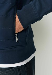 Granatowa bluza z zamkiem błyskawicznym, z teksturowanymi panelami, ściągaczami na rękawach oraz kieszenią na zamek. Noszona na białej koszuli, w połączeniu z ciemnymi dżinsami.
