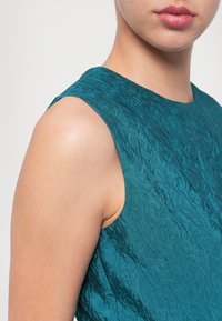 Rochie fără mâneci de culoare teal, confecționată dintr-un material texturat, creponat. Apropierea arată un gât larg și un umăr neted, subliniind materialul unic.